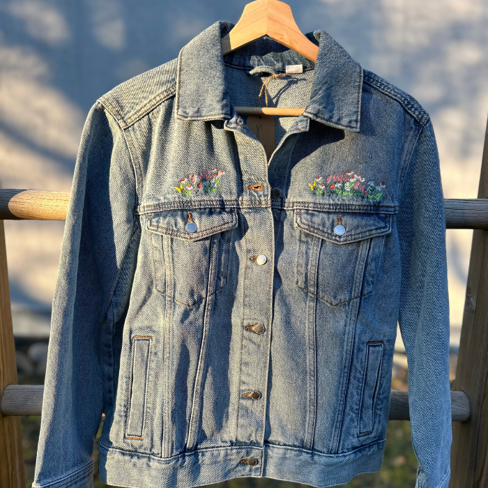 Denim jacket with floral embroidery on a wooden hanger against a blurred natural background