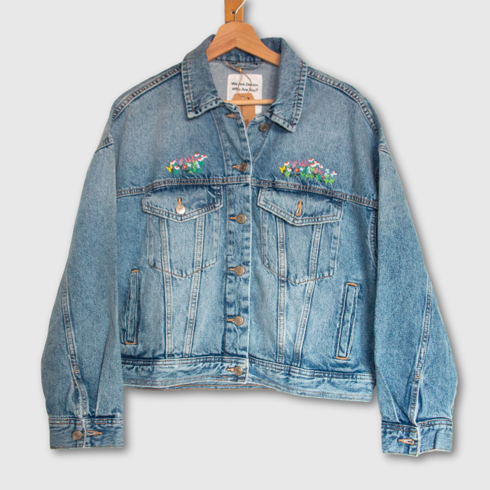 Blue denim jacket with floral embroidery on a hanger against a light gray background