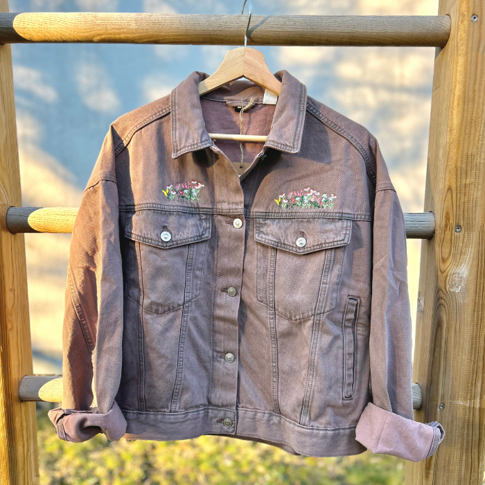 Washed Pink denim jacket with floral embroidery on a wooden railing against a blurred natural background